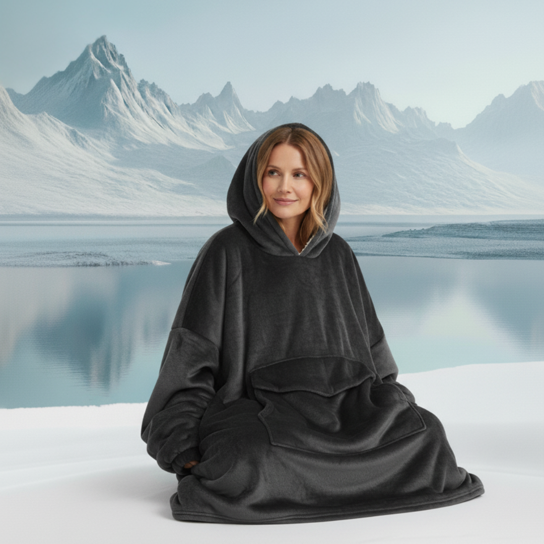 Person wearing a black hooded blanket in a snowy landscape with mountains and water.