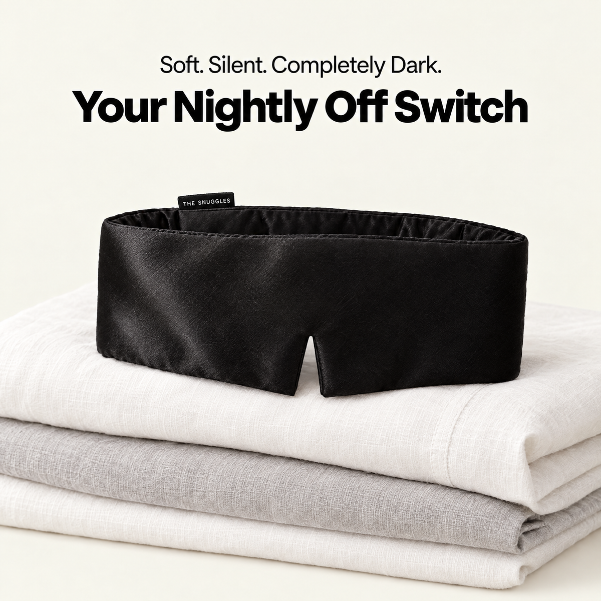 The Snuggles Silky Sleep Mask resting on folded linen bedding