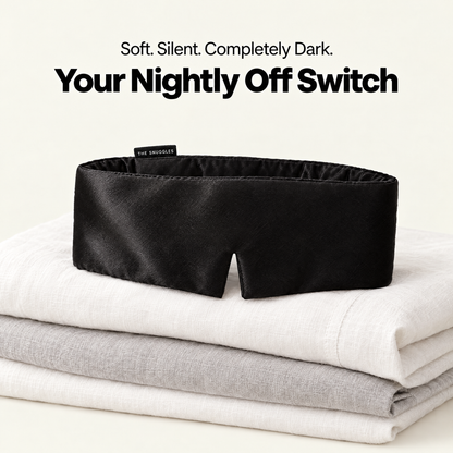 The Snuggles Silky Sleep Mask resting on folded linen bedding